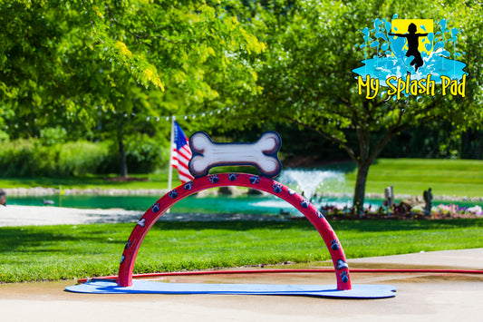 Doggy Wash Water Play Feature by My Portable Splash Pad – red arch with blue paw prints and white bone design installed on outdoor concrete with grass and trees in background.