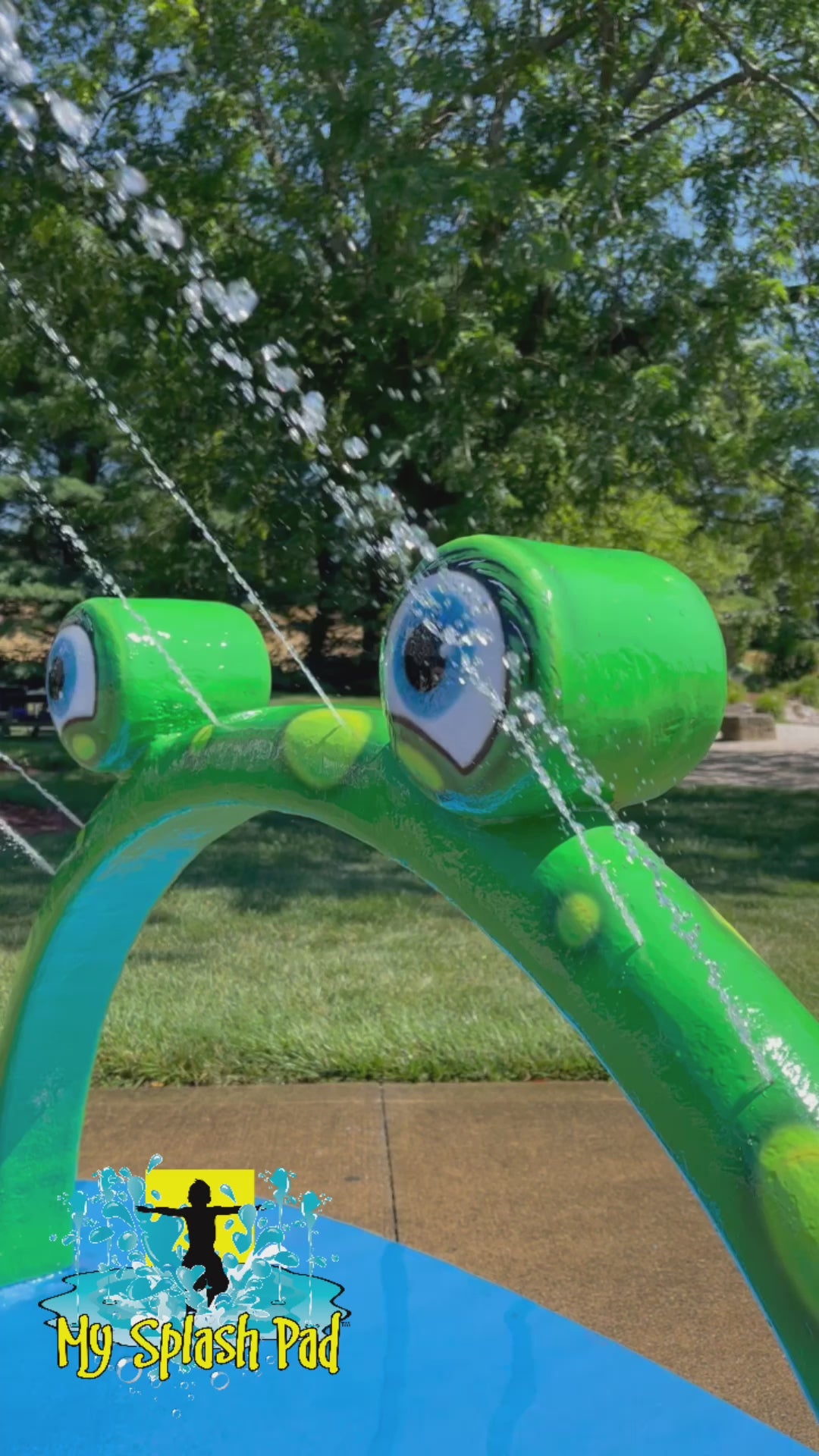 Mini Frog Arch Mobile Water Play Feature by My Portable Splash Pad – green arch with frog eyes installed on outdoor concrete with grass, trees, and American flag in background.