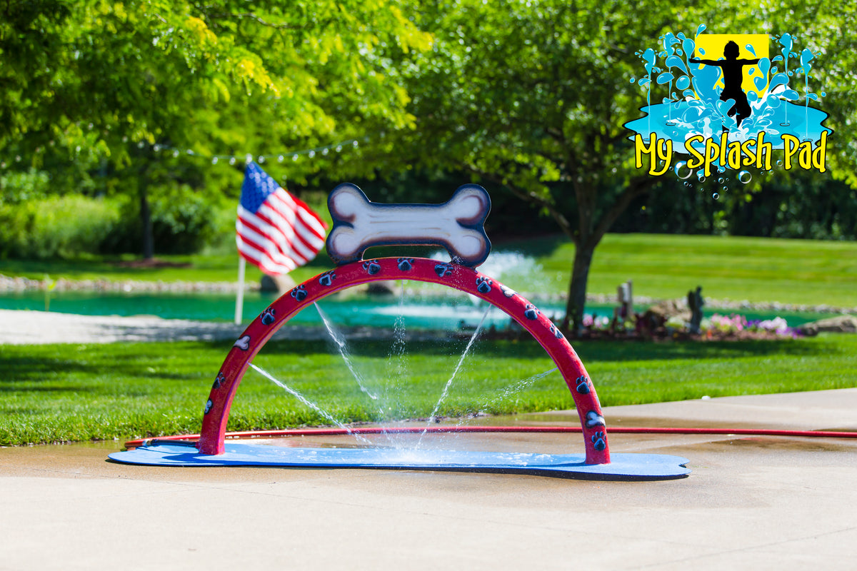 Doggy Wash Water Play Feature by My Portable Splash Pad – red arch with blue paw prints and white bone spraying water streams on outdoor concrete with grass, trees, and American flag in background.