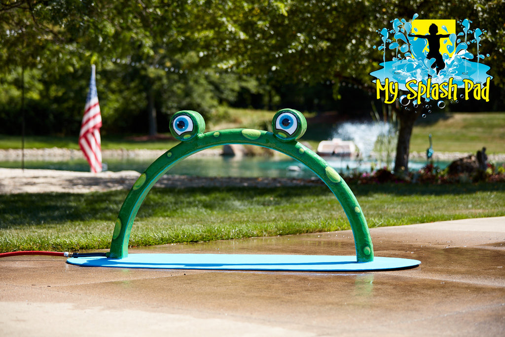 Mini Frog Arch Mobile Water Play Feature by My Portable Splash Pad – green arch with frog eyes installed on outdoor concrete with grass, trees, and American flag in background.