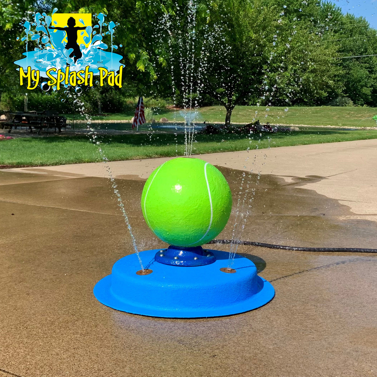 A portable splash pad featuring a green tennis ball at the top, with water spraying from multiple nozzles on a blue fiberglass base, placed on a concrete surface outdoors.